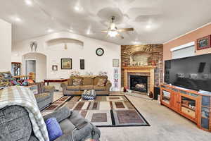 Carpeted living room with vaulted ceiling, a stone fireplace, and ceiling fan