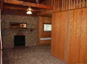 Unfurnished living room featuring wood walls, a stone fireplace, ceiling fan, baseboard heating, and carpet floors