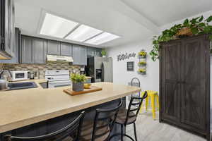 Kitchen with gray cabinetry, white electric range, sink, stainless steel fridge with ice dispenser, and a breakfast bar area