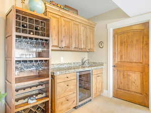 Bar with light carpet, light stone counters, sink, light brown cabinets, and wine cooler