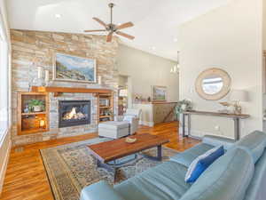 Living room with ceiling fan with notable chandelier, light wood-type flooring, vaulted ceiling, and a stone fireplace