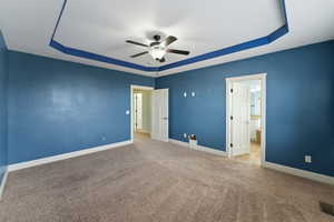 Unfurnished bedroom with light colored carpet, a raised ceiling, ensuite bath, and ceiling fan