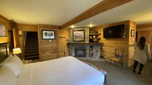 Carpeted bedroom featuring beam ceiling, wooden walls, and a fireplace