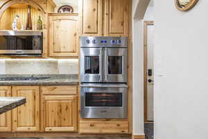 Kitchen with dark stone countertops, decorative backsplash, light brown cabinets, and appliances with stainless steel finishes