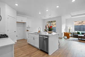 Kitchen with stainless steel dishwasher, sink, light hardwood / wood-style flooring, white cabinetry, and an island with sink