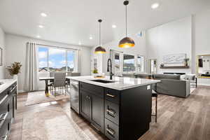 Kitchen featuring sink, hanging light fixtures, light stone counters, stainless steel dishwasher, and a center island with sink