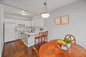 Kitchen with kitchen peninsula, backsplash, white appliances, white cabinetry, and hanging light fixtures