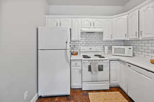 Kitchen with white cabinets, dark hardwood / wood-style flooring, white appliances, and tasteful backsplash