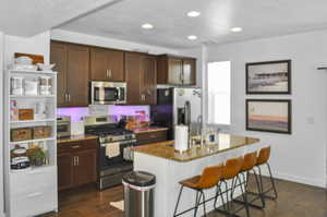 Kitchen with a kitchen bar, light stone counters, stainless steel appliances, dark hardwood / wood-style floors, and an island with sink