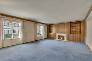 Unfurnished living room with wooden walls, built in shelves, ornamental molding, a fireplace, and carpet floors