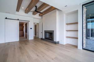 Unfurnished living room with ceiling fan, a barn door, light hardwood / wood-style floors, beam ceiling, and a large fireplace