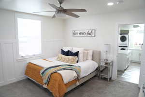 Bedroom featuring dark carpet, stacked washing maching and dryer, ensuite bath, and ceiling fan