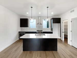 Kitchen with wall chimney exhaust hood, tasteful backsplash, an island with sink, pendant lighting, and appliances with stainless steel finishes