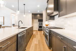 Kitchen featuring sink, wall chimney exhaust hood, light wood-type flooring, appliances with stainless steel finishes, and decorative light fixtures