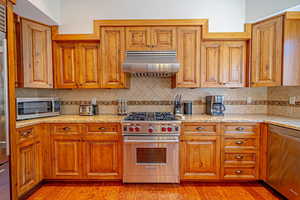 Kitchen featuring light stone countertops, ventilation hood, decorative backsplash, appliances with stainless steel finishes, and light wood-type flooring