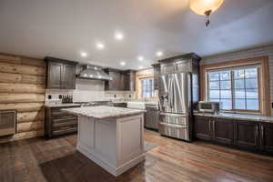 Kitchen featuring appliances with stainless steel finishes, dark hardwood / wood-style flooring, rustic walls, wall chimney range hood, and a center island