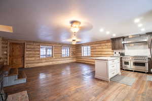 Kitchen featuring wall chimney exhaust hood, white cabinetry, log walls, and double oven range