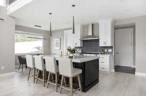 Kitchen featuring white cabinetry, wall chimney range hood, light hardwood / wood-style flooring, an island with sink, and pendant lighting