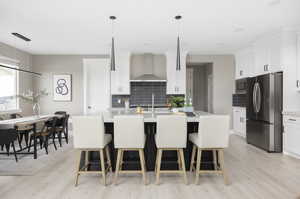 Kitchen featuring wall chimney exhaust hood, hanging light fixtures, stainless steel appliances, decorative backsplash, and white cabinets