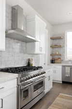 Kitchen featuring backsplash, dark wood-type flooring, wall chimney exhaust hood, appliances with stainless steel finishes, and white cabinetry