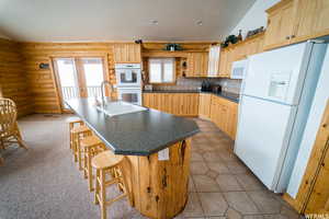 Kitchen with light brown cabinets, white appliances, a center island with sink, and rustic walls