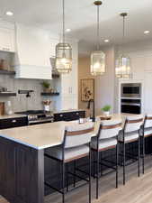 Kitchen with light wood-type flooring, custom range hood, stainless steel appliances, a center island with sink, and white cabinetry