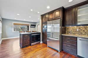 Kitchen featuring decorative backsplash, light stone countertops, light wood-type flooring, dark brown cabinetry, and stainless steel appliances