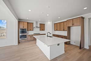 Kitchen with stainless steel double oven, wall chimney range hood, gas stovetop, a spacious island, and light wood-type flooring