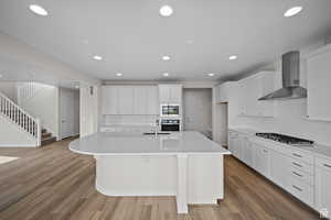 Kitchen with white cabinetry, stainless steel appliances, wall chimney range hood, an island with sink, and wood-type flooring