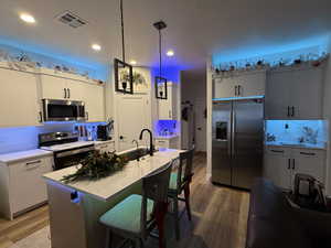 Kitchen featuring a kitchen bar, appliances with stainless steel finishes, a kitchen island with sink, wood-type flooring, and decorative light fixtures