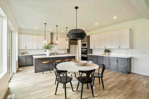 Kitchen featuring white cabinetry, wall chimney range hood, an island with sink, pendant lighting, and gray cabinets