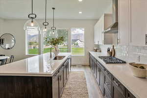 Kitchen with stainless steel gas stovetop, sink, hanging light fixtures, wall chimney exhaust hood, and an island with sink