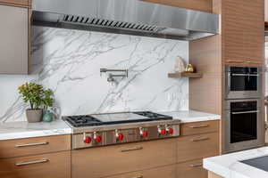 Kitchen featuring wall chimney exhaust hood, light stone counters, stainless steel appliances, and tasteful backsplash