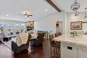 Living room with dark wood-type flooring, french doors, ceiling fan with notable chandelier, vaulted ceiling with beams, and a fireplace