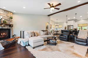 Living room featuring vaulted ceiling with beams, ceiling fan with notable chandelier, a stone fireplace, and dark wood-type flooring