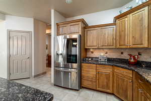 Kitchen featuring decorative backsplash, light tile patterned flooring, stainless steel fridge with ice dispenser, and dark stone counters