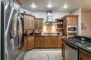 Kitchen with dark stone countertops, wall chimney range hood, appliances with stainless steel finishes, and tasteful backsplash