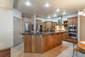 Kitchen with decorative backsplash, dark stone counters, wall chimney exhaust hood, stainless steel appliances, and a center island with sink