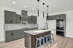 Kitchen with built in microwave, sink, light stone counters, a center island with sink, and custom range hood