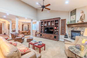 Living room featuring light carpet, decorative columns, ceiling fan with notable chandelier, vaulted ceiling, and a stone fireplace