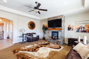 Carpeted living room featuring a tray ceiling, a stone fireplace, and ceiling fan