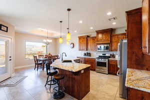 Kitchen featuring appliances with stainless steel finishes, a kitchen breakfast bar, light stone counters, a center island with sink, and a notable chandelier