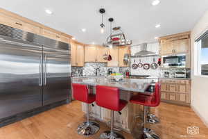 Kitchen featuring light stone counters, sink, wall chimney range hood, built in appliances, and a center island