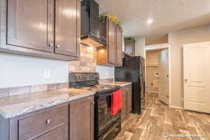 Kitchen with light stone countertops, wall chimney exhaust hood, a textured ceiling, black appliances, and light hardwood / wood-style flooring