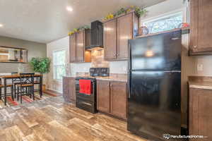 Kitchen featuring black appliances, wall chimney range hood, a textured ceiling, tasteful backsplash, and light hardwood / wood-style floors