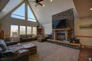 Living room featuring ceiling fan, dark carpet, a stone fireplace, and high vaulted ceiling