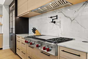 Kitchen with stainless steel gas cooktop, premium range hood, light stone countertops, light brown cabinets, and backsplash