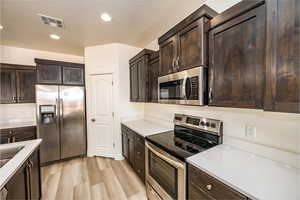 Kitchen with a textured ceiling, light hardwood / wood-style flooring, stainless steel appliances, and dark brown cabinets