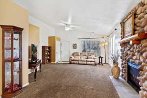 Carpeted living room featuring a textured ceiling, a stone fireplace, ceiling fan, and vaulted ceiling