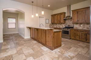 Kitchen with light stone countertops, stainless steel gas range oven, wall chimney exhaust hood, decorative light fixtures, and a breakfast bar area